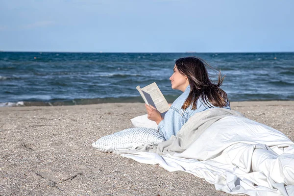 Genç bir kadın sahilde bir battaniyeyle kaplanmış olarak uzanıyor ve bir kitap okuyor. Sahildeki rahat atmosfer, yaz konsepti..