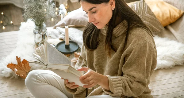 A young woman in a sweater reads a book in a cozy home. The concept of comfort and Hobbies.