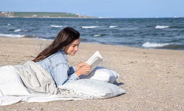 Genç bir kadın sahilde bir battaniyeyle kaplanmış olarak uzanıyor ve bir kitap okuyor. Sahildeki rahat atmosfer, yaz konsepti..