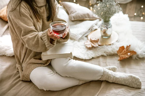 Cozy autumn or winter at home, a woman in a knitted sweater with a Cup of tea and a book .