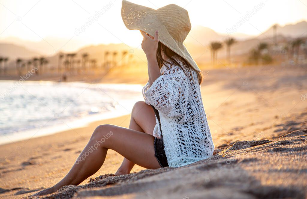 Beautiful boho model in a swimsuit with a white Cape with a hat on her head poses on the beach at sunset