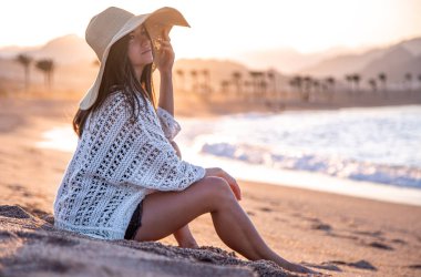 Güneşin batışında, kafasında şapka olan beyaz bir mayo giymiş güzel bir manken kumsalda poz veriyor.