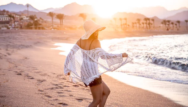 A beautiful boho model in a white Cape and swimsuit poses on the beach in the sunlight
