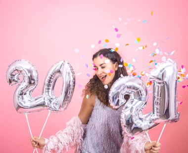 Beautiful young brunette girl with curly hair and festive clothes posing on a pink studio background with confetti on her face and holding in her hand silver balloons from the numbers 2021 clipart