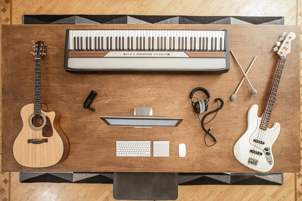 Composition of acoustic guitar, bass guitar, musical keys, a man at the computer and headphones and a shelf for drums on a large wooden table.