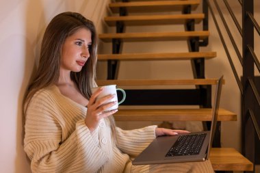 Side portrait of a young woman holding a mug and watching the laptop screen in soft evening light. Home comfort and focus.