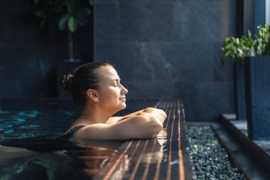A woman rests on the warm pool edge with eyes closed as sunlight falls on her face. Soft reflections and clean lines evoke wellness, mindfulness and a personal pause.