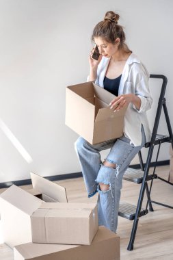 A beautiful young woman sits on a stepladder with an open box while talking on the phone. Focused yet slightly excited mood, daylight, cozy minimalism and real-life vibe.