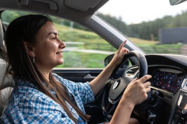 A woman in a plaid shirt smiles while driving a car, enjoying the road and the travel experience.