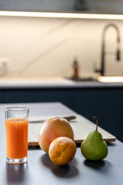 Parlak bir havuç suyu, portakal armudu ve greyfurtla birlikte koyu bir tezgahın üzerinde duruyor. Yumuşak LED ışıklandırması, sıcacık minimalist bir hava katıyor..