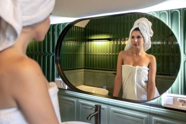 One young woman in a white towel looks at her reflection in a large oval mirror against green tiles. Home spa, morning routine and clean aesthetic with a calm fresh lifestyle vibe.