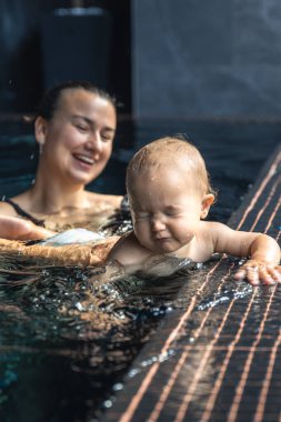 Bebek suya dokunur ve anne yakınlarda otururken, su sıçrattığı için şaşı kalır. Işık ve yansımalar samimi bir keşif ve rahatlatıcı bir aile refahı hissi yakalar..