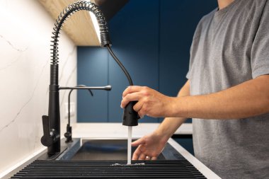 Diğer el suyu, mavi dolapları ve mat siyah musluğu kontrol ederken, diğeri de her gün düzenli enerjili spreyleri hedef alan bir adamın yan açısı..