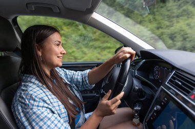 Kareli tişörtlü, gezintide araba süren, yolun ve anın tadını çıkaran gülümseyen kadın. Sakin atmosfer, modern yaşam tarzı ve günlük yaşam tarzı.