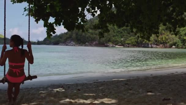 Una joven en traje de baño, balanceándose en un columpio de cuerda hecho en casa, en una playa de arena en la laguna azul . — Vídeo de stock