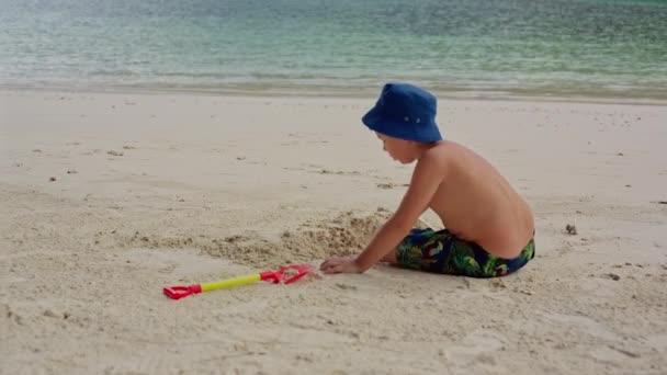 Dziecko siedzi na plaży w pobliżu wody i wykopuje otwór z łopata. — Wideo stockowe