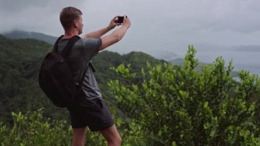 Sırtında sırt çantası olan bir gezgin, bir tepenin tepesinde duruyor ve cep telefonuyla bir manzara çekiyor..