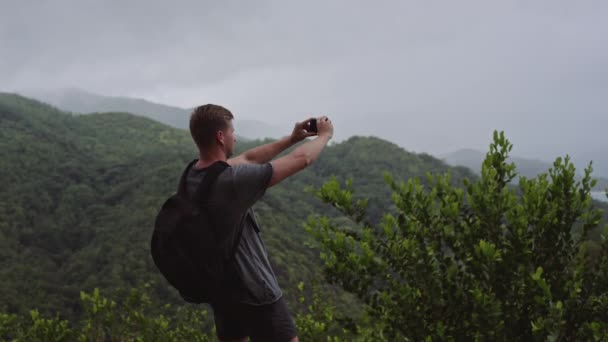 Een reiziger met een rugzak op zijn rug, staat op de top van een heuvel en schiet een landschap op zijn mobiele telefoon. — Stockvideo