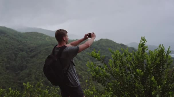 Um viajante com uma mochila nas costas, fica no topo de uma colina e atira em uma paisagem em seu telefone celular . — Vídeo de Stock