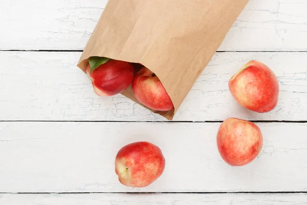 Ripe red peaches in the paper bag on the white wooden table, top view