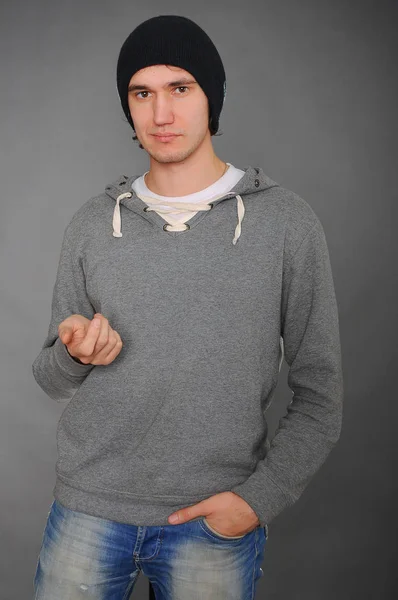 Portrait of an attractive young male model in black hat, jeans and gray sweatshirt, studio, dark gray background