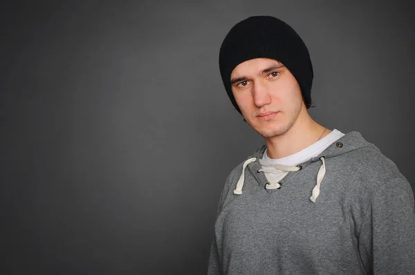Portrait of an attractive young male model in black hat, jeans and gray sweatshirt, studio, dark gray background