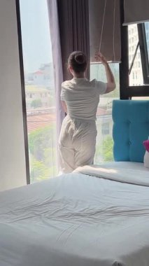 Beyazlı genç kız otelde perdeleri açıyor. Bütün güneş ışığı eve giriyor. Saçlarını açıyor, güzel kestane saçlarını düzeltiyor. .