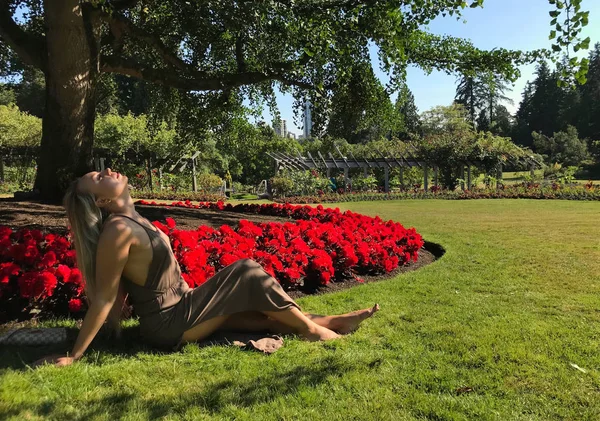 Young woman on vacation in rose garden in Stanley Park. Pretty woman sitting on grass by flower bed in sunny day.  Vancouver. British Columbia. Canada.