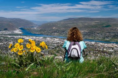 Woman with backpack sitting on the top of mountain enjoying scenic views of city and river. Spring wild flowers on hills. Wenatchee. Washington. United States clipart