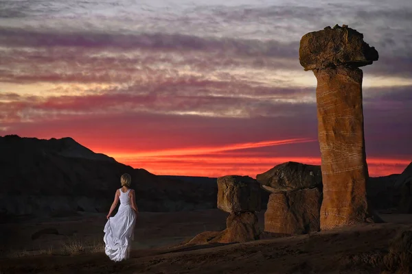 Mujer rubia en vestido blanco por los taburetes gigantes de roca viendo la puesta de sol en el desierto de roca en Arizona . — Foto de Stock