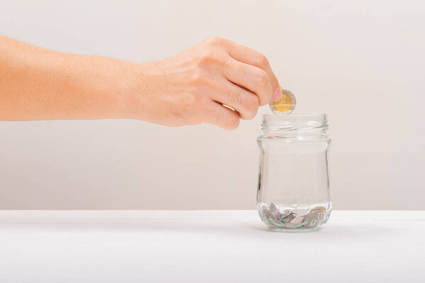 Белый ручной человек держит монету положить в банку или стеклянную бутылку для экономии на мягком белом фоне. Экономия денег, концепция экономии