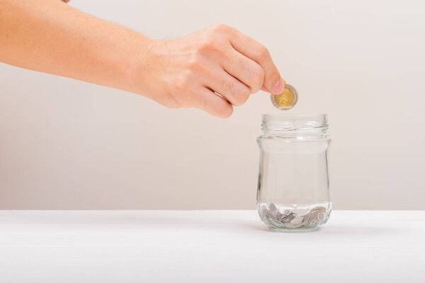 Белый ручной человек держит монету положить в банку или стеклянную бутылку для экономии на мягком белом фоне. Экономия денег, концепция экономии