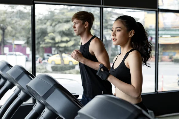 Genç kadın ve erkek spor salonundaki modern elektrik koşu bantlarında yan yana koşuyorlar. Koşu bandında koşan güzel Asyalı kadın ve arka planda zinde bir genç adam. Genç çift elektrikli koşu bandında.