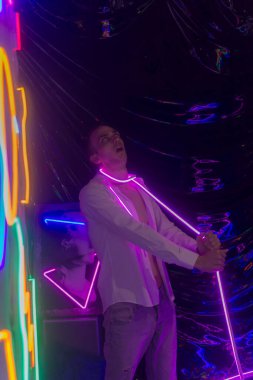 Düğmeleri iliklenmemiş beyaz gömlekli yakışıklı adamın portresi. Neon renkli oda. Eski 90 'lar eski moda. Erkeğe yakın neon kelimelerin yansıması.