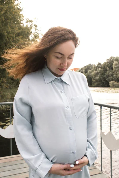Πορτρέτο μιας ευτυχισμένης και περήφανης εγκύου δίπλα στο ποτάμι της πόλης. Γυναίκα με ανοιχτό μπλε πουκάμισο. — Φωτογραφία Αρχείου