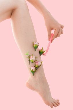 Kadının ince tıraşsız bacağı ve pembe arka planda pembe çiçekler. Kadın bacağını pembe bir jiletle tıraş ediyor..