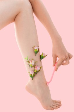 Kadının ince tıraşsız bacağı ve pembe arka planda pembe çiçekler. Kadın bacağını pembe bir jiletle tıraş ediyor..