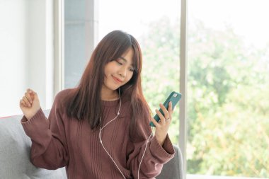 Güzel Asyalı kadın oturma odasındaki gri koltukta akıllı telefon kullanıyor.