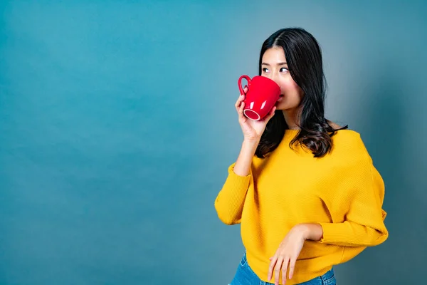Young asian woman in yellow sweater holding a red cup of coffee, smell good and enjoy the coffee on blue background