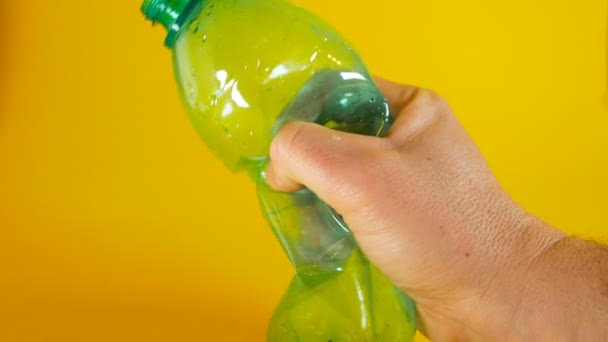 Una mano uomo schiaccia una bottiglia di plastica verde usa e getta su uno sfondo giallo, filmato ideale per temi come l'ecologia e il riciclaggio — Video Stock