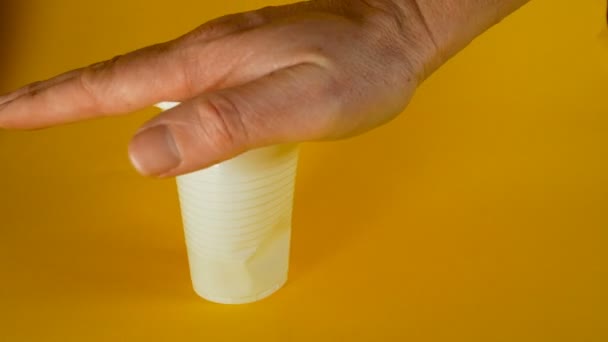 Una mano uomo schiaccia un bicchiere di plastica monouso bianco su uno sfondo giallo, filmato ideale per temi come l'ecologia e il riciclaggio — Video Stock