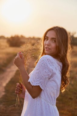 Uzun beyaz elbiseli kız gün batımında gün batımında yol kenarında duran yaz güneşine seviniyor..