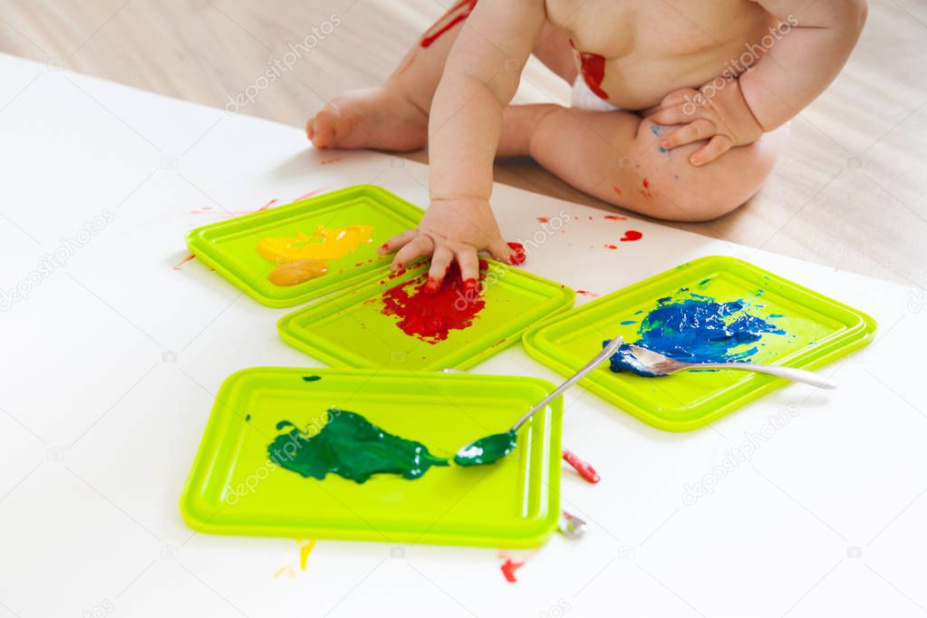 Baby child draws with colored paints hands, dirty feet. White li