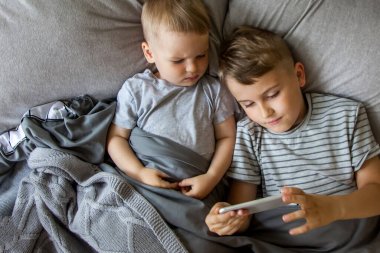 Kahverengi saçlı iki küçük çocuk büyük bir yatakta yatıyor. Gül, telefonda ve tablette çizgi film izle. Cihazı çalıştır. Gri yatak. Çocuklar Kafkasyalı..