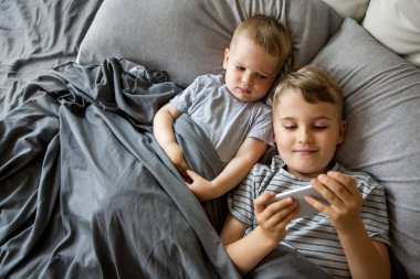 Kahverengi saçlı iki küçük çocuk büyük bir yatakta yatıyor. Gül, telefonda ve tablette çizgi film izle. Cihazı çalıştır. Gri yatak. Çocuklar Kafkasyalı..