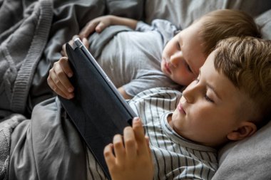 Kahverengi saçlı iki küçük çocuk büyük bir yatakta yatıyor. Gül, telefonda ve tablette çizgi film izle. Cihazı çalıştır. Gri yatak. Çocuklar Kafkasyalı..