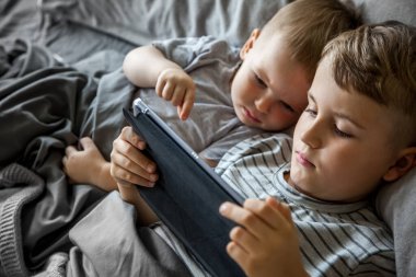 Kahverengi saçlı iki küçük çocuk büyük bir yatakta yatıyor. Gül, telefonda ve tablette çizgi film izle. Cihazı çalıştır. Gri yatak. Çocuklar Kafkasyalı..