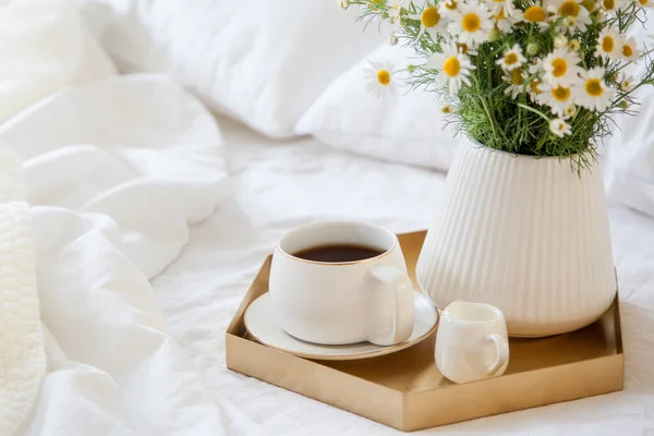 Morning breakfast in bed. White bedding. Golden tray in the nordic style. White cup with black coffee and a milkman with milk. Daisies flowers in a vase. White little alarm clock
