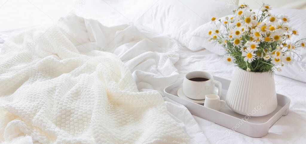 Morning breakfast in bed. White bedding. Golden tray in the nordic style. White cup with black coffee and a milkman with milk. Daisies flowers in a vase. White little alarm clock