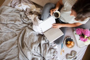 Gri yatakta servis edilen kruvasan ve kahveyle kahvaltı. Güzel genç kız yatakta uzanıyor ve kitap okuyor..